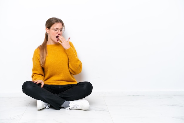 Jeune femme caucasienne assise sur le sol isolé sur fond blanc bâillant et couvrant la bouche grande ouverte avec la main