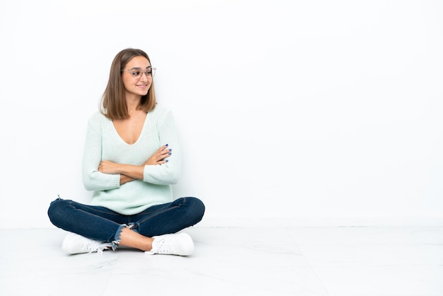 Jeune femme caucasienne assise sur le sol isolé sur fond blanc avec les bras croisés et heureux