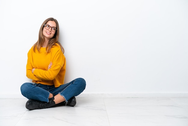 Jeune femme caucasienne assise sur le sol isolé sur fond blanc avec les bras croisés et heureux