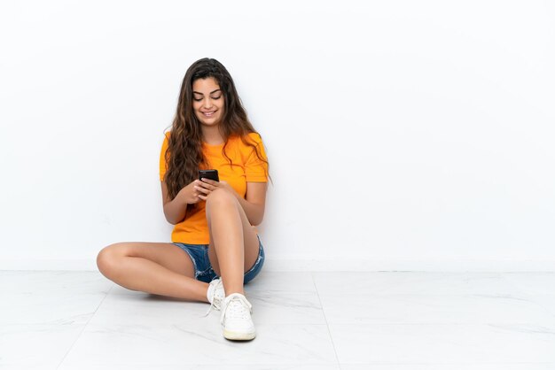 Jeune femme caucasienne assise sur le sol isolé sur fond blanc envoyant un message avec le mobile