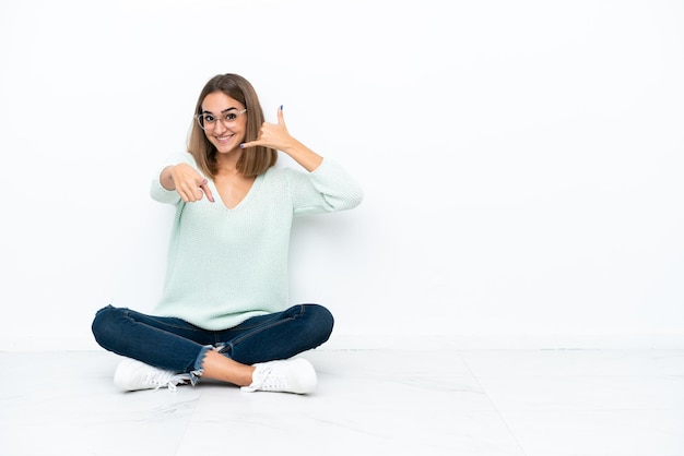 Jeune femme caucasienne assise sur le sol isolé sur fond blanc faisant un geste de téléphone et pointant vers l'avant