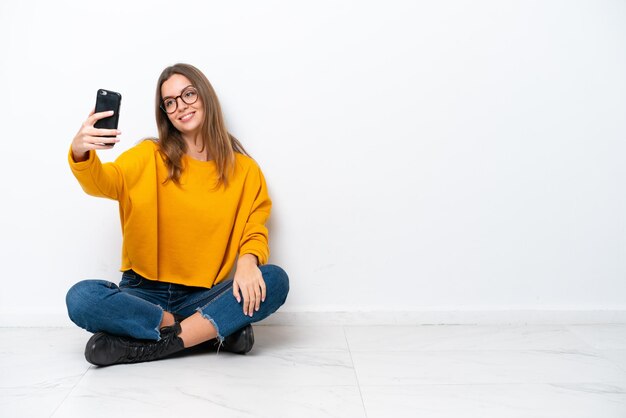 Jeune femme caucasienne assise sur le sol isolé sur fond blanc faisant un selfie