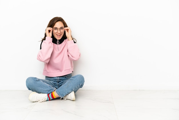 Jeune femme caucasienne assise sur le sol isolé sur fond blanc avec des lunettes et surpris