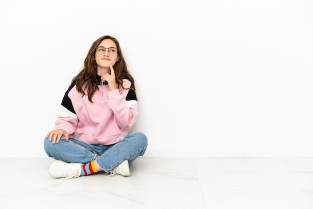 Jeune femme caucasienne assise sur le sol isolé sur fond blanc en pensant à une idée tout en levant les yeux