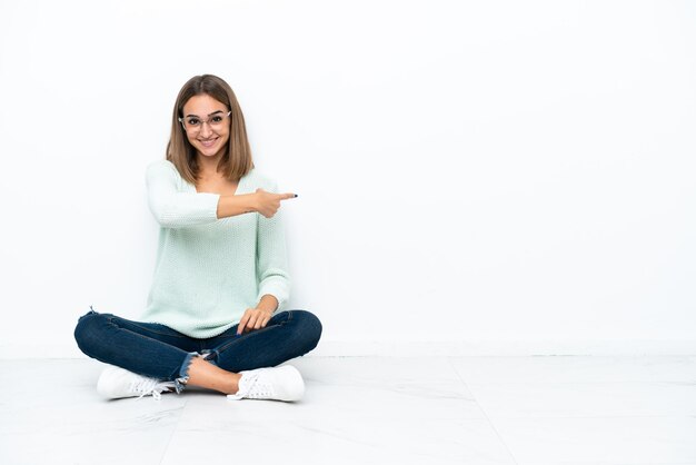 Jeune femme caucasienne assise sur le sol isolé sur fond blanc pointant vers l'arrière