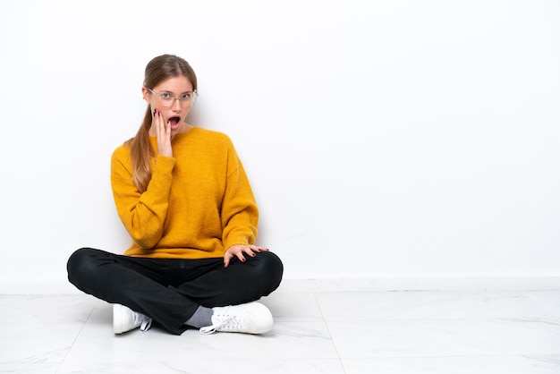 Jeune femme caucasienne assise sur le sol isolé sur fond blanc surpris et choqué en regardant à droite