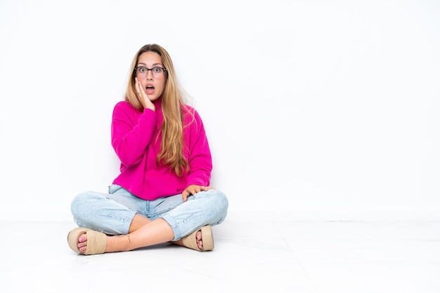 Jeune femme caucasienne assise sur le sol isolé sur fond blanc surpris et choqué en regardant à droite