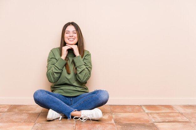 Jeune femme caucasienne assise sur le sol isolé garde les mains sous le menton, regarde joyeusement de côté.