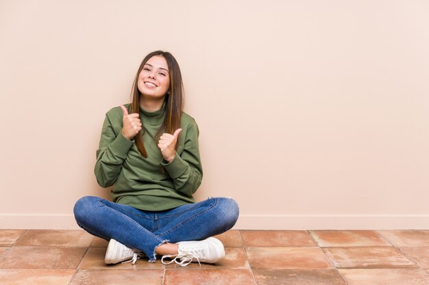 Jeune femme caucasienne assise sur le sol isolé en levant les deux pouces vers le haut, souriant et confiant.