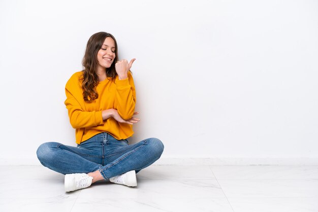 Jeune femme caucasienne assise sur le sol isolé sur un mur blanc pointant vers le côté pour présenter un produit