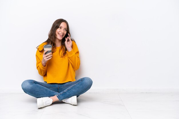 Jeune femme caucasienne assise sur le sol isolé sur un mur blanc tenant du café à emporter et un mobile