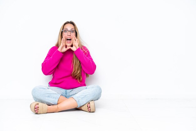 Jeune femme caucasienne assise sur le sol isolée sur fond blanc criant et annonçant quelque chose