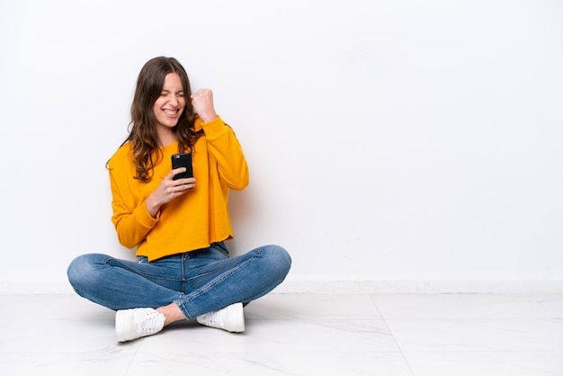 Jeune femme caucasienne assise sur le sol isolée sur un mur blanc avec téléphone en position de victoire