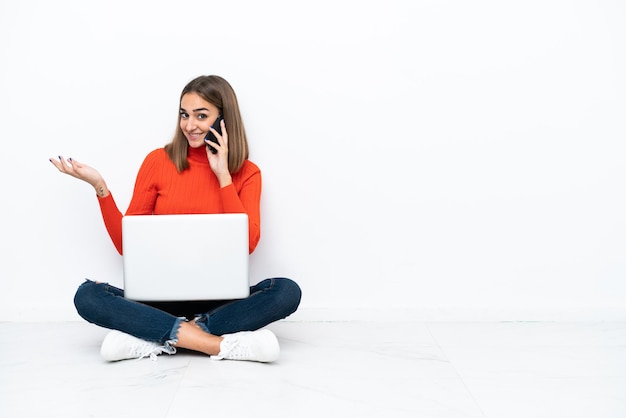 Jeune femme caucasienne assise sur le sol avec un ordinateur portable en gardant une conversation avec le téléphone portable avec quelqu'un