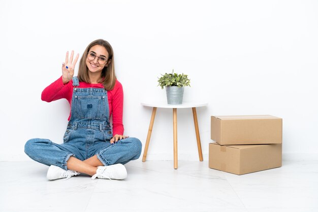 Jeune femme caucasienne assise sur le sol parmi des boîtes heureuses et comptant trois avec les doigts