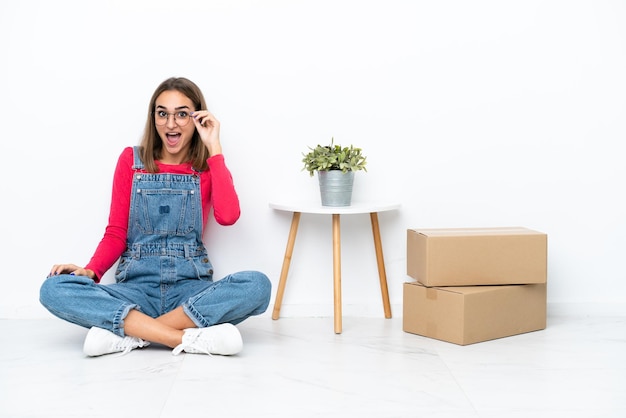 Jeune femme caucasienne assise sur le sol parmi des boîtes avec des lunettes et surprise