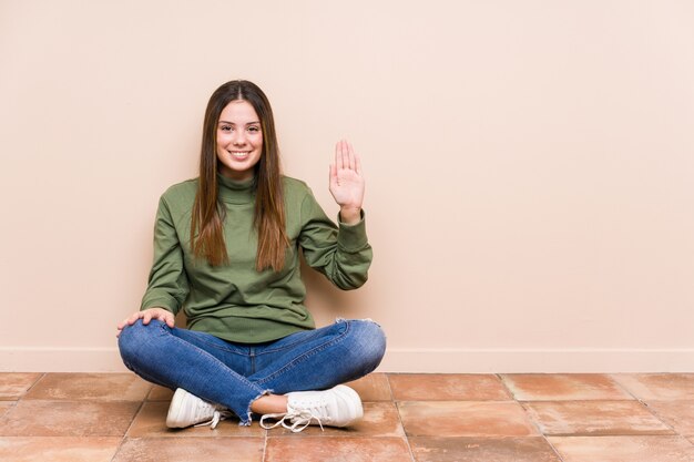 Jeune femme caucasienne assise sur le sol souriant joyeux montrant le numéro cinq avec les doigts.