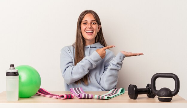 Jeune femme caucasienne assise à une table avec un équipement de sport isolé sur fond blanc excité tenant un espace de copie sur la paume.