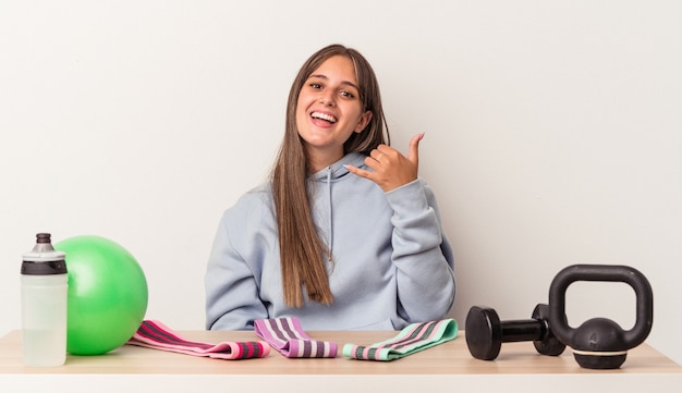 Jeune femme caucasienne assise à une table avec un équipement de sport isolé sur fond blanc montrant un geste d'appel de téléphone portable avec les doigts.