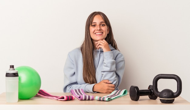 Jeune femme caucasienne assise à une table avec un équipement de sport isolé sur fond blanc souriant heureux et confiant, touchant le menton avec la main.