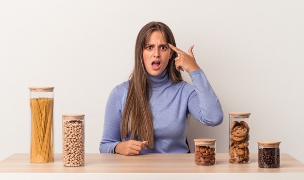 Jeune femme caucasienne assise à une table avec un pot de nourriture isolé sur fond blanc montrant un geste de déception avec l'index.