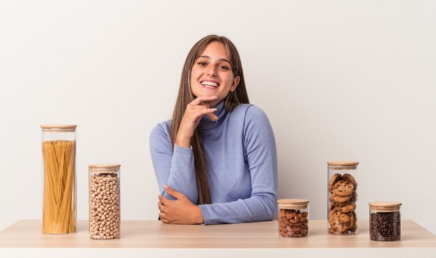 Jeune Femme Caucasienne Assise à Une Table Avec Un Pot De Nourriture Isolé Sur Fond Blanc Souriant Heureux Et Confiant, Touchant Le Menton Avec La Main.