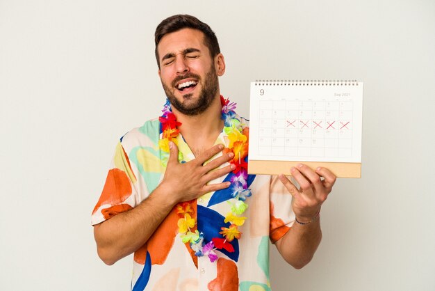 Jeune femme caucasienne attendant ses vacances tenant un calendrier isolé sur fond blanc éclate de rire en gardant la main sur la poitrine.