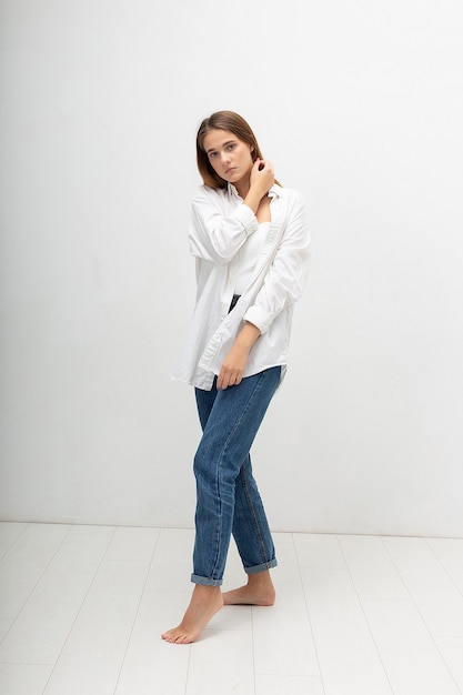 Jeune Femme Caucasienne Attirante Avec De Longs Cheveux Bruns En Chemise, Un Jean Bleu Sur Fond De Studio Blanc. Jolie Dame Maigre Posant Pieds Nus Dans Une Pièce Lumineuse. Portrait De Belle Femme