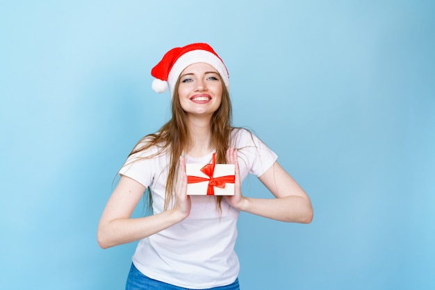 Jeune femme caucasienne au chapeau de Noël et avec des cadeaux en t-shirt blanc sourit de bonheur avec...