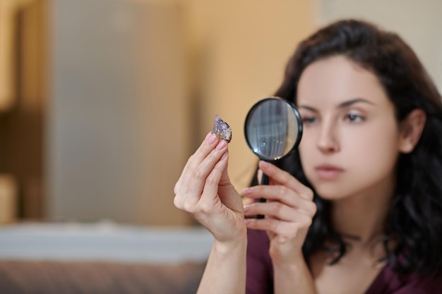 Jeune femme caucasienne aux cheveux noirs et concentrée examinant une améthyste dans sa main à l'aide d'une loupe