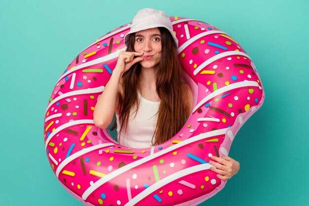 Jeune femme caucasienne avec un beignet gonflable isolé sur fond bleu avec les doigts sur les lèvres gardant un secret.