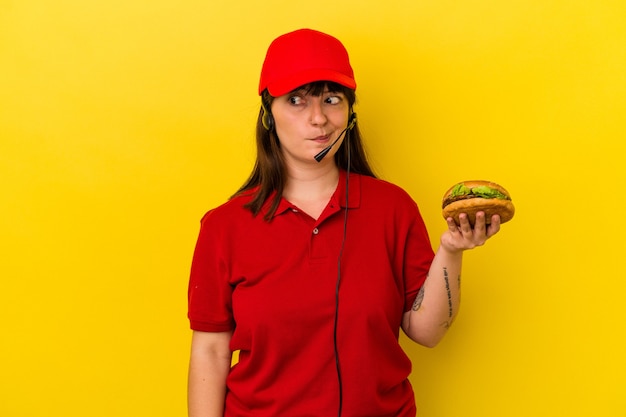 Photo jeune femme caucasienne bien roulée tenant un hamburger isolé sur fond jaune confus, se sent dubitative et incertaine.