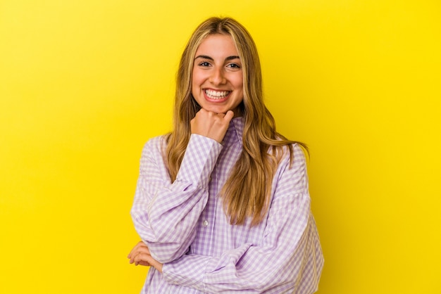 Jeune femme caucasienne blonde isolée sur fond jaune souriant heureux et confiant, toucher le menton avec la main.