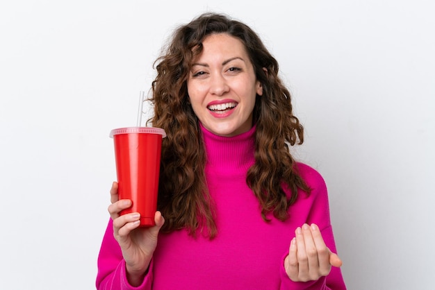 Jeune femme caucasienne buvant du soda isolé sur fond blanc invitant à venir avec la main Heureux que vous soyez venu