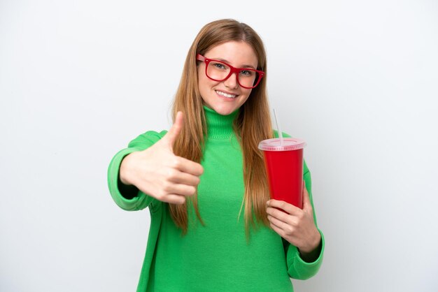 Jeune femme caucasienne buvant du soda isolé sur fond blanc avec les pouces vers le haut parce que quelque chose de bien s'est produit