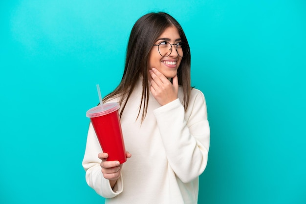 Jeune femme caucasienne buvant du soda isolé sur fond bleu regardant sur le côté et souriant