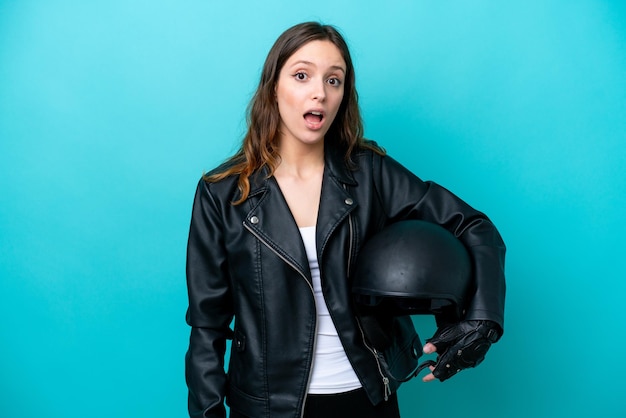 Jeune femme caucasienne avec un casque de moto isolé sur fond bleu avec une expression faciale surprise