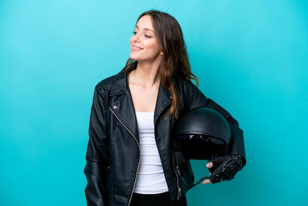 Jeune femme caucasienne avec un casque de moto isolé sur fond bleu regardant sur le côté et souriant