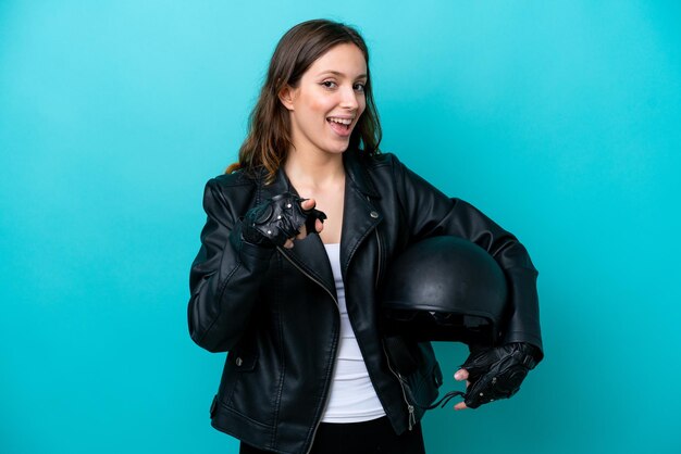 Jeune femme caucasienne avec un casque de moto isolé sur fond bleu surpris et pointant vers l'avant