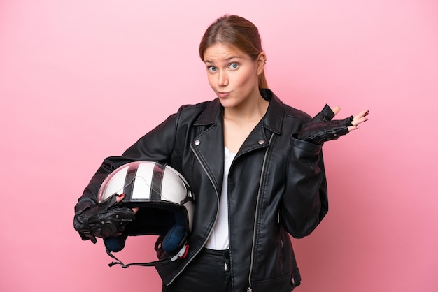 Jeune femme caucasienne avec un casque de moto isolé sur fond rose ayant des doutes tout en levant les mains