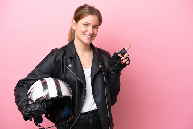 Jeune femme caucasienne avec un casque de moto isolé sur fond rose pointant le doigt sur le côté