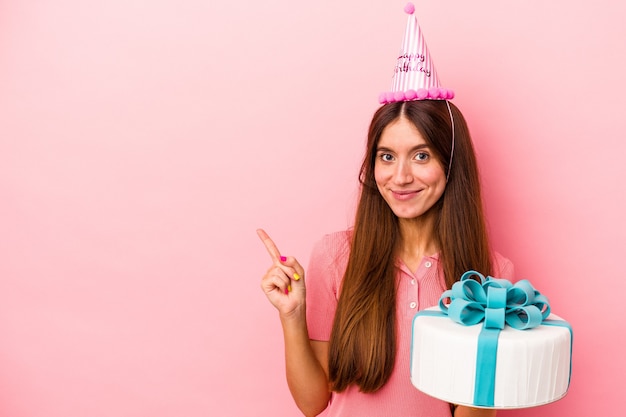 Jeune femme caucasienne célébrant un anniversaire isolé sur fond rose souriant et pointant de côté, montrant quelque chose dans un espace vide.