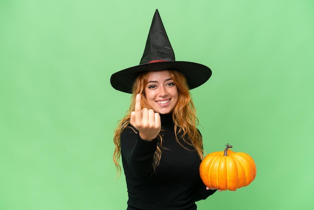 Jeune femme caucasienne costume comme sorcière tenant une citrouille isolée sur fond vert chroma key faisant un geste à venir