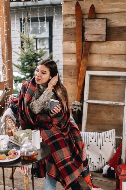 Jeune femme caucasienne dans un plaid boit une boisson chaude sur la véranda avec arbre de Noël et décorations.