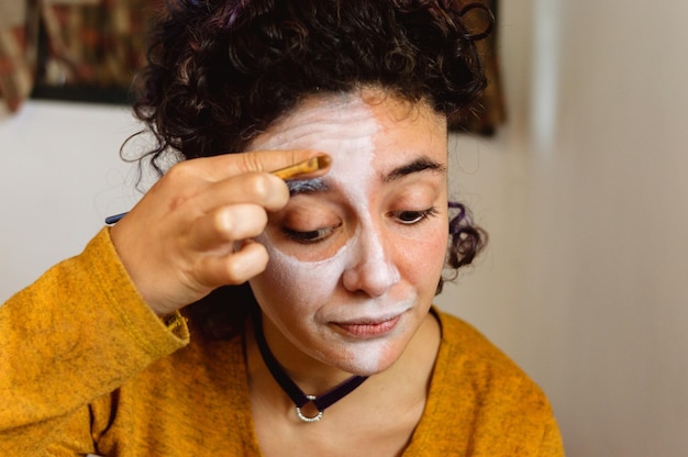 La jeune femme caucasienne dans sa chambre applique le maquillage artistique blanc avec une petite brosse