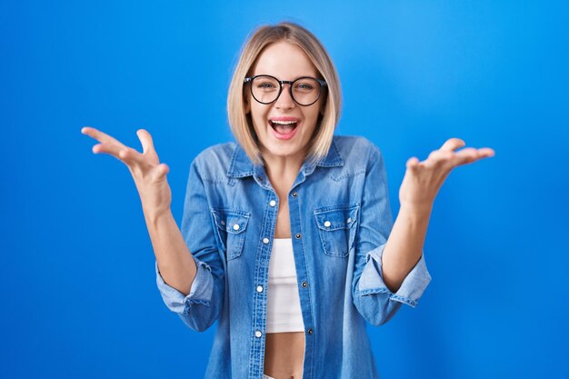 Jeune femme caucasienne debout sur fond bleu célébrant la folie et étonnée du succès avec les bras levés et les yeux ouverts hurlant d'excitation. notion de gagnant