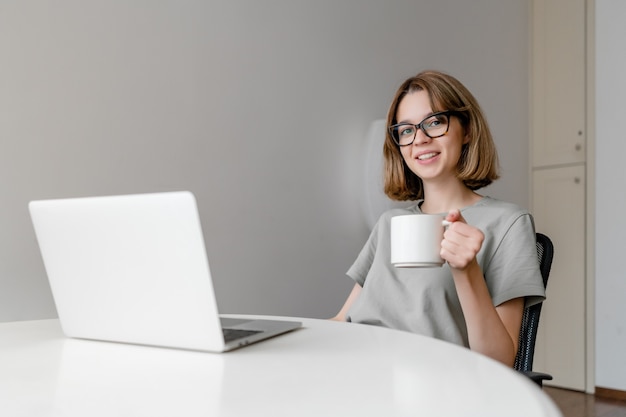 Jeune femme caucasienne détendue souriant assis avec un ordinateur portable dans l'appartement avec une tasse dans ses mains