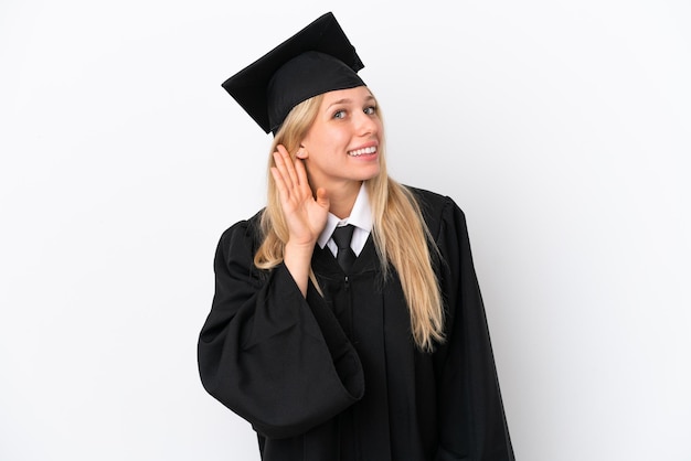 Jeune femme caucasienne diplômée universitaire isolée sur fond blanc écoutant quelque chose en mettant la main sur l'oreille