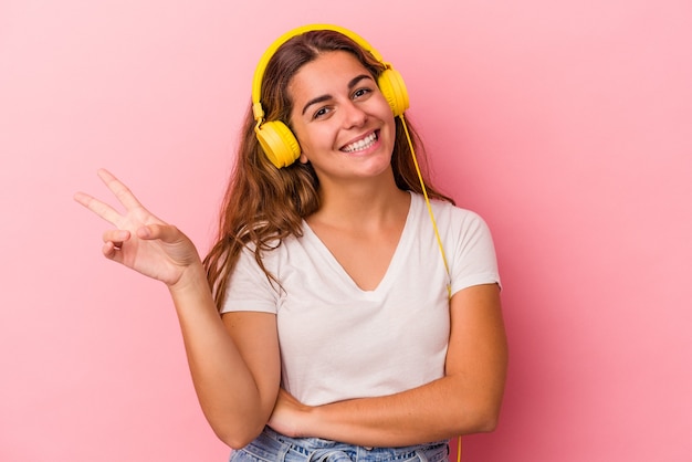 Jeune femme caucasienne écoutant de la musique isolée sur fond rose joyeuse et insouciante montrant un symbole de paix avec les doigts.