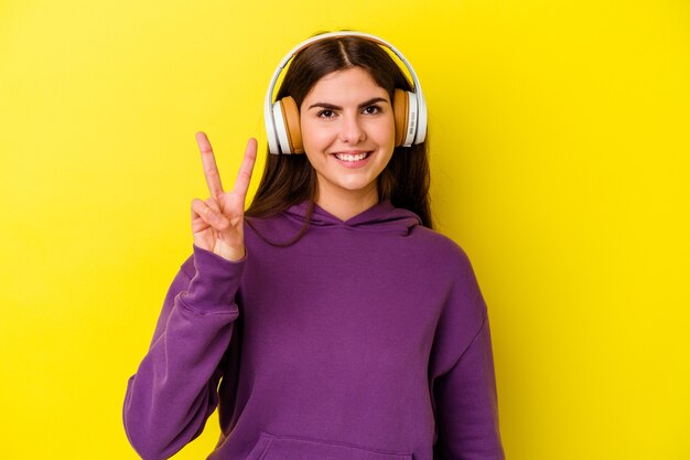 Jeune femme caucasienne, écouter de la musique avec un casque isolé sur un mur rose montrant le numéro deux avec les doigts.